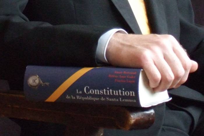 Eine Hand liegt auf einem Buch mit dem Titel "La Constitution de la République de Santa Lemusa".