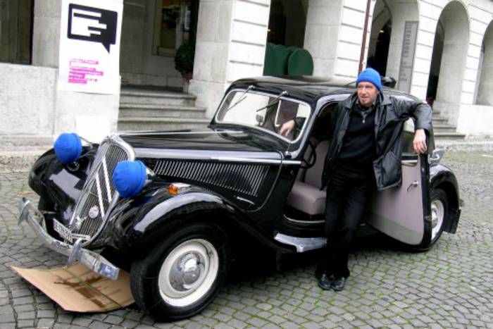 Ein altes Auto mit blauen Kappen über den Scheinwerfern, Fahrer mit ebensolcher Kappe.